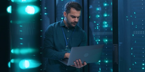 An IT professional using a laptop computer with rack server cabinets behind him.